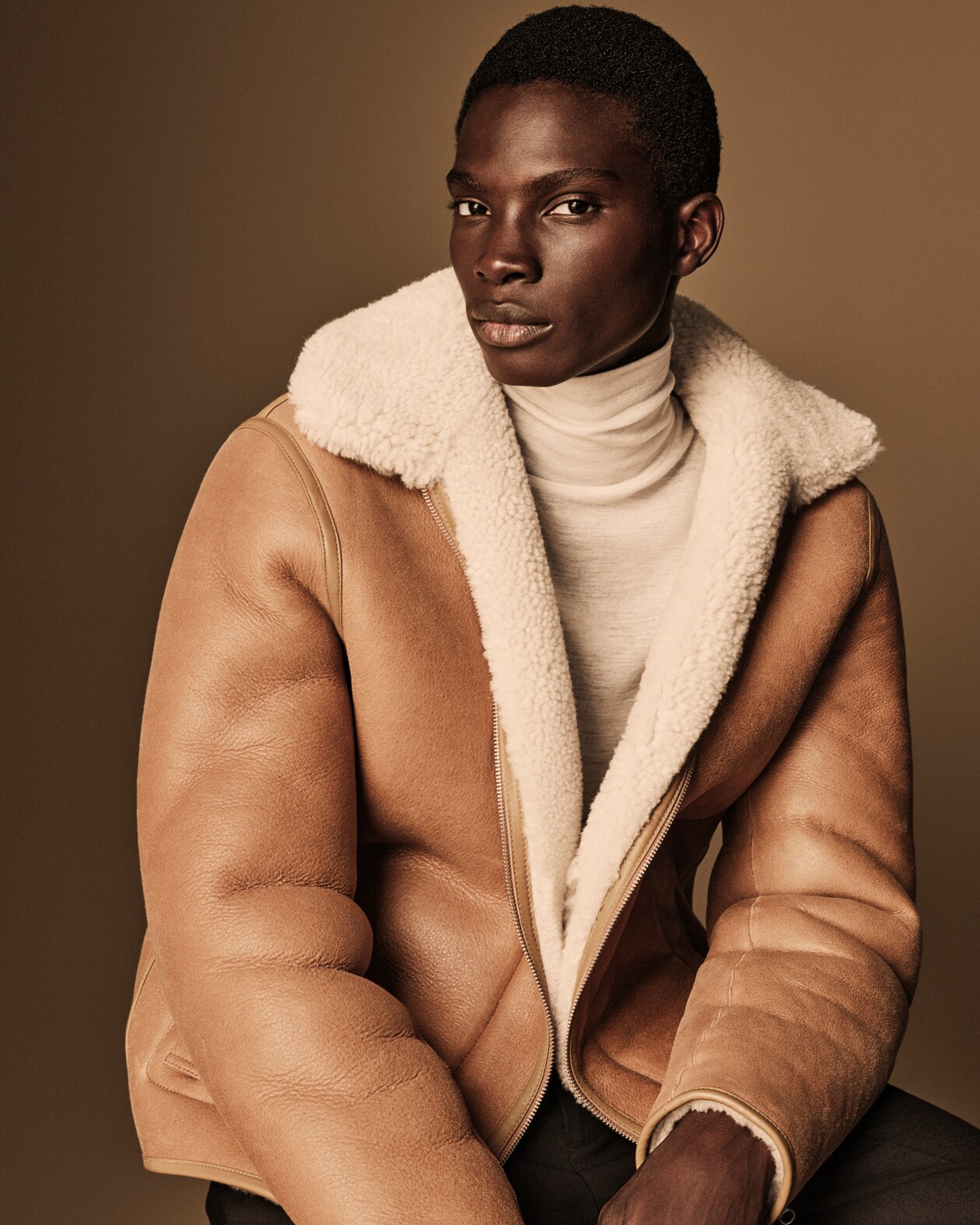 Man wears a brown leather shearling jacket layered over a white turtleneck top